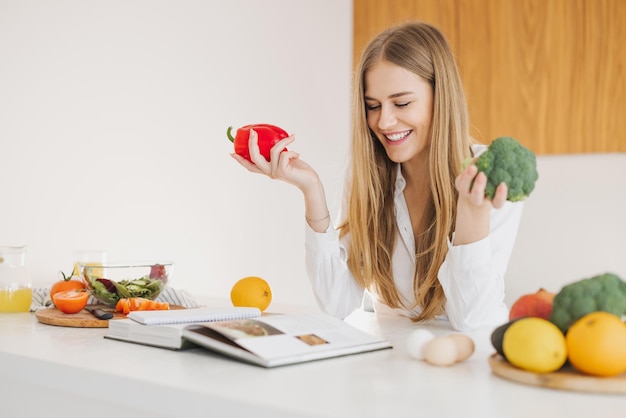 Retrato de uma linda garota loira sorridente segurando pimentão e brócolis e lendo o livro de receitas na cozinha na mesa entre cozinhar ingredientes