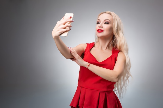 Retrato de uma linda garota sorridente bem sucedida fazendo selfie em um vestido vermelho sobre fundo claro