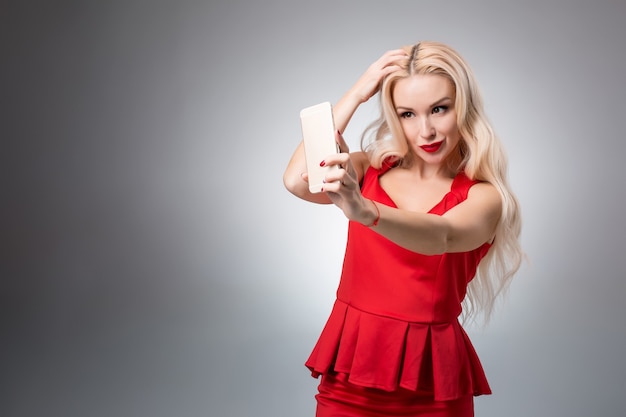 Retrato de uma linda garota sorridente bem sucedida fazendo selfie em um vestido vermelho sobre fundo claro