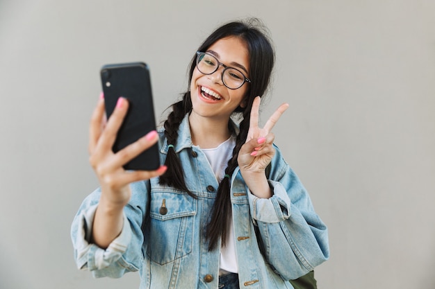 Retrato de uma linda garota sorridente fofa na jaqueta jeans, usando óculos isolados sobre uma parede cinza usando telefone celular, tome uma selfie mostrando paz.