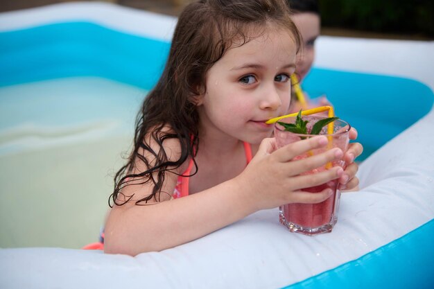 Retrato de uma linda garotinha caucasiana bebendo coquetel na piscina ao ar livre no quintal da casa