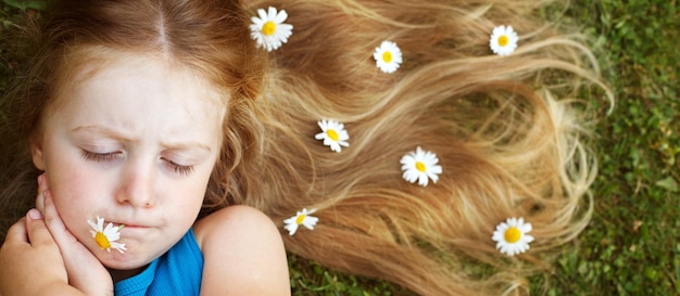 Retrato de uma linda garotinha com cabelo vermelho saudável com flores de camomila deitada no banner de grama