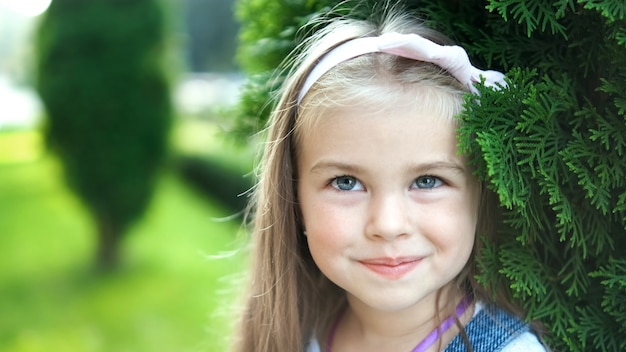 Retrato de uma linda garotinha em pé ao ar livre no parque de verão, sorrindo alegremente.