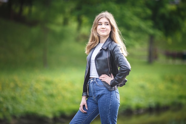 Retrato de uma linda garotinha estilosa em jeans azuis e jaqueta de couro no parque da cidade no fundo da floresta verde