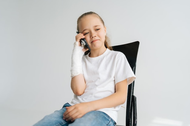 Retrato de uma linda garotinha loira com braço quebrado envolto em bandagem de gesso falando smartphone sentado na cadeira em fundo branco