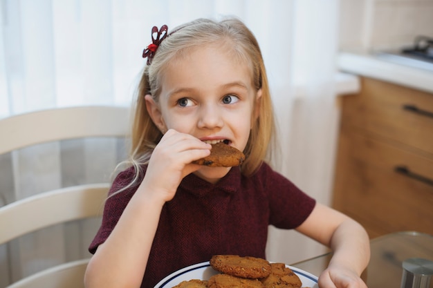 Retrato de uma linda garotinha loira comendo biscoitos de manhã na cozinha.