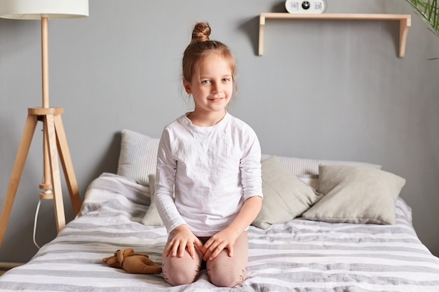 Retrato de uma linda garotinha sorridente com penteado de coque vestindo roupas casuais domésticas sentada na cama olhando para a câmera com emoções positivas manhã tranquila em casa