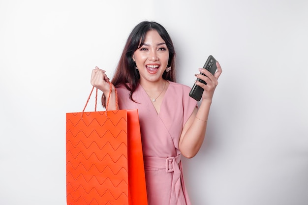 Retrato de uma linda jovem asiática feliz em pé, animada, segurando uma sacola de compras on-line e sua foto de estúdio de smartphone isolada em fundo branco