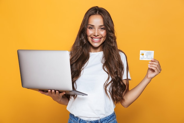 Retrato de uma linda jovem com longos cabelos castanhos em pé sobre uma parede amarela, segurando um laptop, mostrando um cartão de crédito de plástico