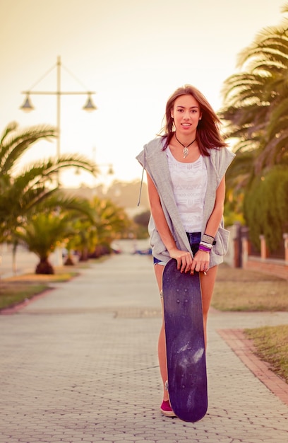 Retrato de uma linda jovem com shorts curtos e skate ao ar livre em um dia quente de verão. Edição de tons quentes.