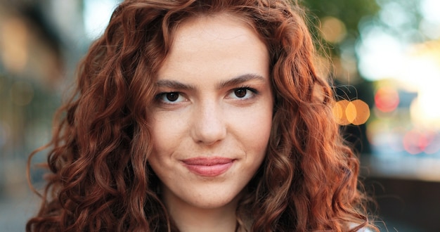 Retrato de uma linda jovem de etnia caucasiana, com cabelo ruivo encaracolado em pé na rua da cidade quente de primavera, vestida com as roupas casuais durante a manhã.