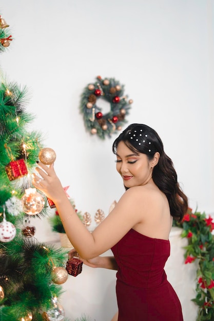 Retrato de uma linda jovem decorando a árvore de Natal sorrindo usando vestido vermelho na sala de estar decorada de Natal dentro de casa