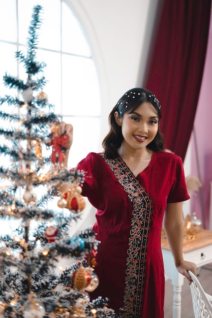 Retrato de uma linda jovem decorando a árvore de Natal sorrindo usando vestido vermelho na sala de estar decorada de Natal dentro de casa