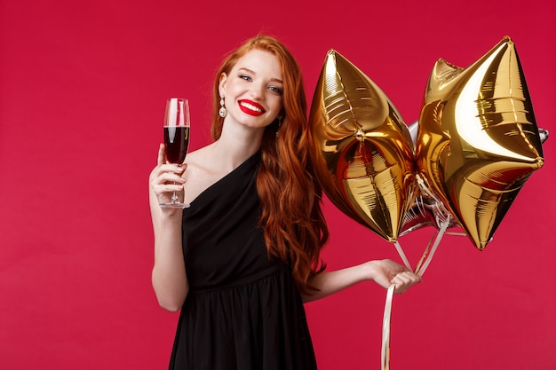 Retrato de uma linda jovem elegante com batom vermelho, cabelo ruivo e vestido preto, segurando balões e taça de champanhe, dizendo brinde para amigo ter aniversário sorrindo alegre