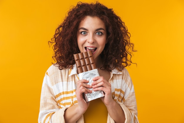 Retrato de uma linda jovem encaracolada bonita posando isolado na parede amarela com chocolate.