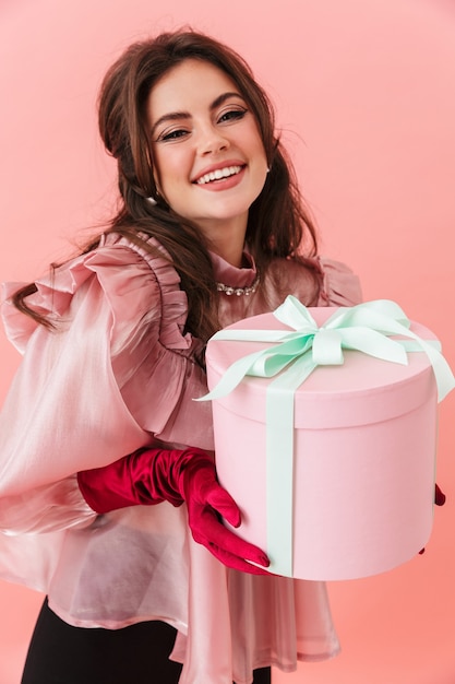 Retrato de uma linda jovem morena alegre com maquiagem brilhante, vestindo roupas da moda em pé isolado na parede rosa, segurando uma caixa de presente