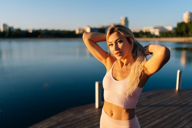 Retrato de uma linda loira esportiva em roupas esportivas em pé posando no píer de madeira no parque cite na manhã ensolarada de verão olhando para a câmera