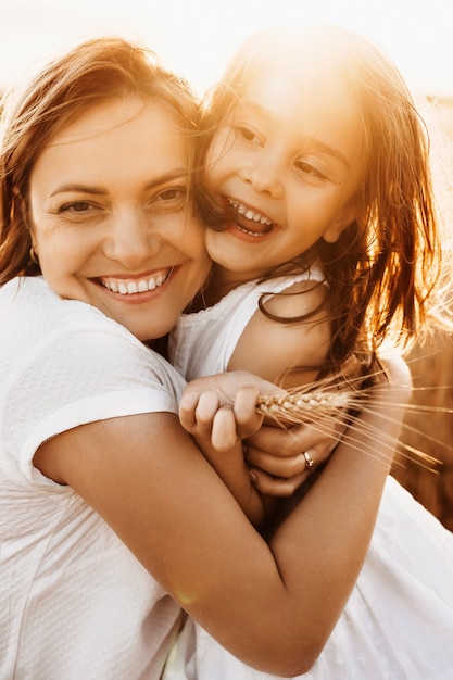 Foto retrato de uma linda mãe abraçando a filha rindo contra o pôr do sol, vestida de branco.