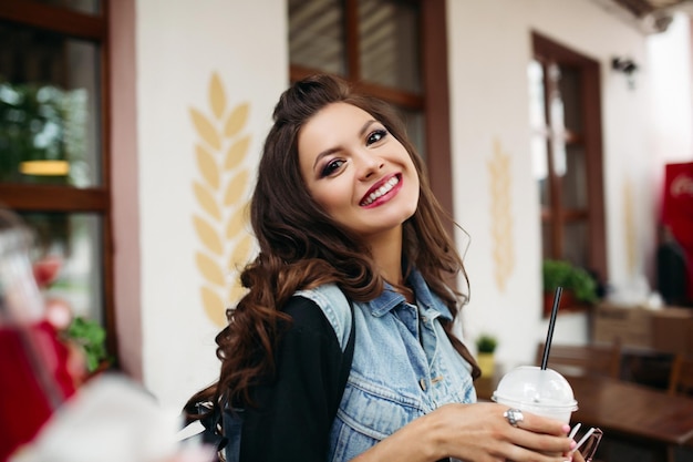 Retrato de uma linda menina com cabelo longo ondulado, sorrindo para a câmera segurando o copo de refrigerante no café.