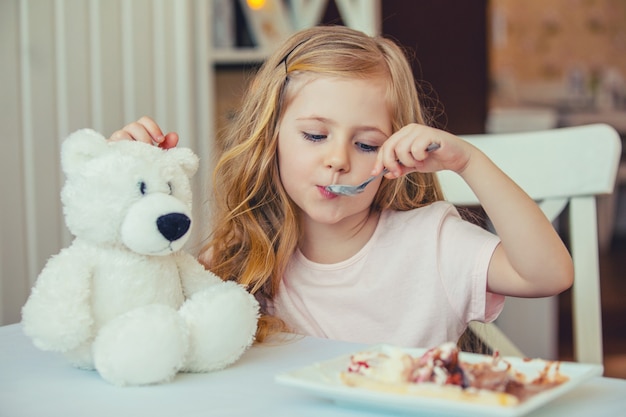 Retrato de uma linda menina com um ursinho de pelúcia em um café, comendo um delicioso sorvete.