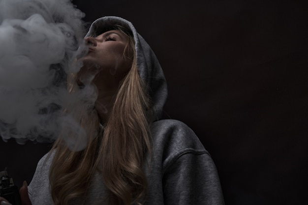 Retrato de uma linda menina loira com capuz cinza fuma vapor isolado no fundo preto do estúdio, nuvem de fumaça, mini cachimbo de água
