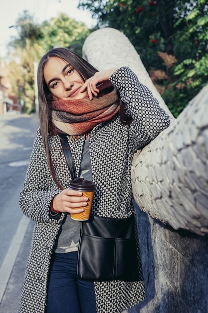 Retrato de uma linda menina morena com bebida para viagem na rua. lenço e casaco de outono na cidade. Vista incrível da mulher de negócios com uma xícara de café, descendo a rua da cidade.