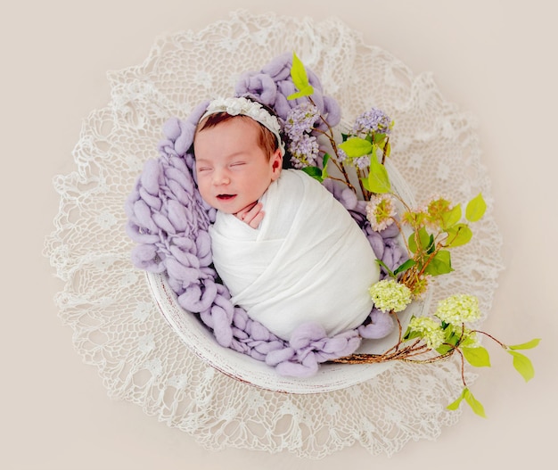 Retrato de uma linda menina recém-nascida enrolada em tecido e usando coroa de flores com flores dormindo e sorrindo na bacia com decoração durante a sessão de fotos de estúdio. Linda criança infantil