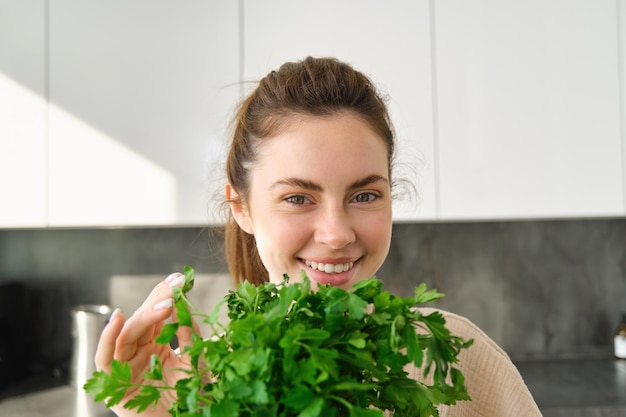 Retrato de uma linda menina sorridente com buquê de salsa em pé na cozinha e cozinhando