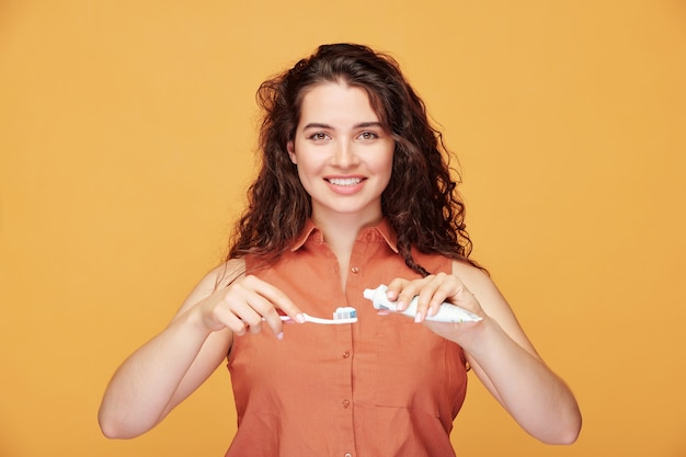 Retrato de uma linda menina sorridente com cabelo encaracolado, aplicando pasta de dente antes de escovar os dentes em laranja