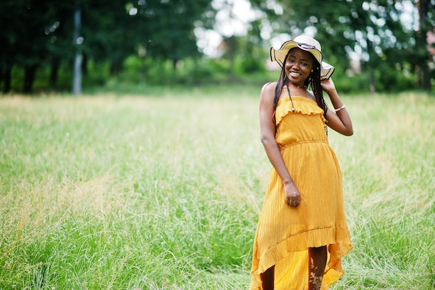 Retrato de uma linda mulher afro-americana de 20 anos com vestido amarelo e chapéu de verão posando na grama verde no parque.