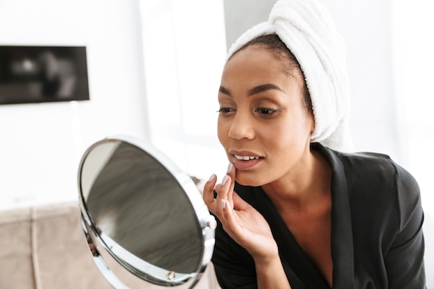 Retrato de uma linda mulher afro-americana em roupão, enrolada em uma toalha branca, aplicando creme facial contra o espelho