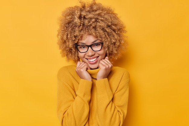 Retrato de uma linda mulher alegre com cabelo espesso encaracolado mantém as mãos na gola do jumper usa óculos transparentes feliz em conhecê-lo posa sobre fundo amarelo Conceito de emoções positivas