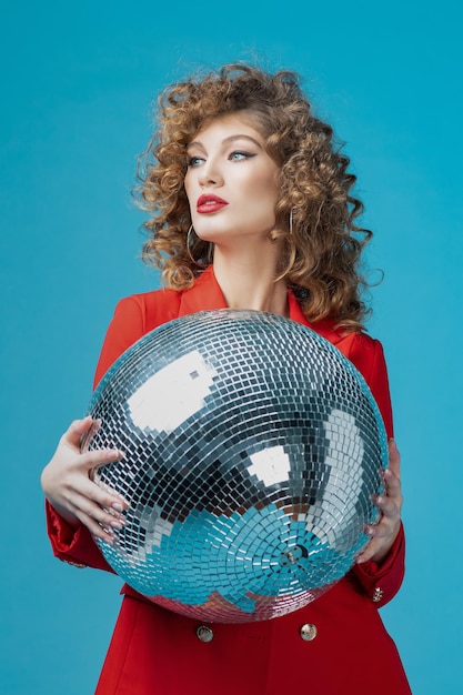 Retrato de uma linda mulher alegre de vestido vermelho fazendo uma festa em pé com bola de discoteca isolada sobre fundo azul