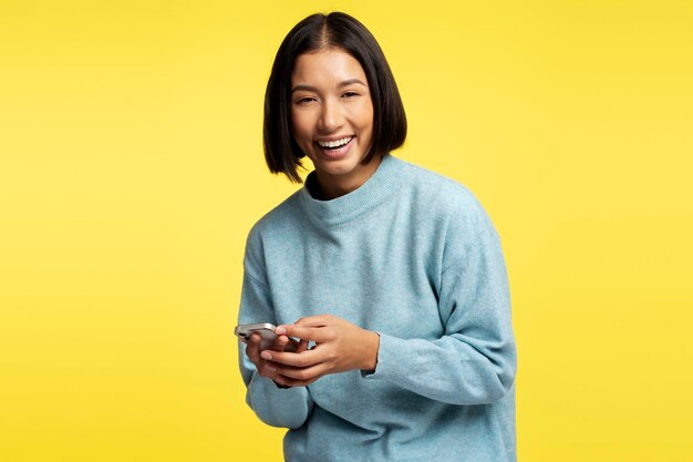 Foto retrato de uma linda mulher asiática sorridente segurando um telefone móvel usando um aplicativo móvel olhando para a câmera de pé isolada em fundo amarelo conceito de compras on-line