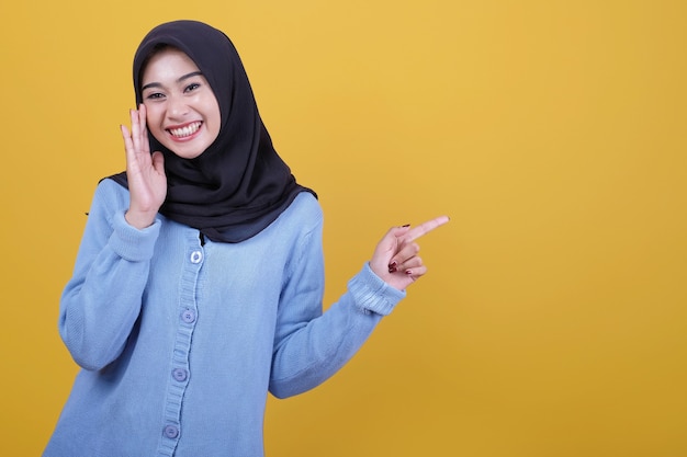 Retrato de uma linda mulher asiática usando hijab preto, dizendo algo sussurrado e apontando com o dedo indicador