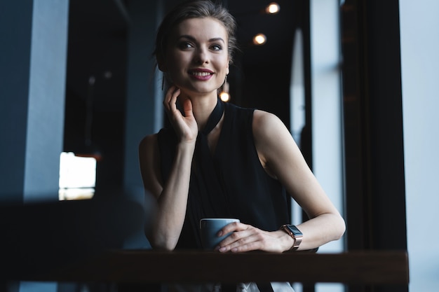 Retrato de uma linda mulher bebendo chá ou café e olhando com sorriso para fora da janela da cafeteria, enquanto desfruta de seu tempo de lazer, almoço de mulher de negócios em um café moderno durante o intervalo do trabalho.