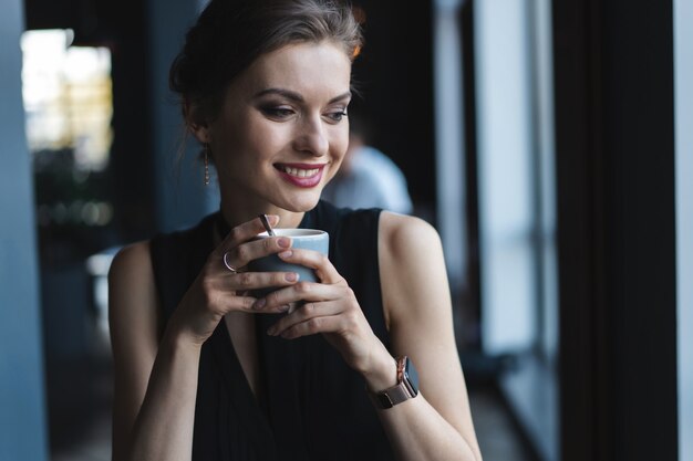 Retrato de uma linda mulher bebendo chá ou café e olhando com sorriso para fora da janela da cafeteria, enquanto desfruta de seu tempo de lazer, almoço de mulher de negócios em um café moderno durante o intervalo do trabalho.