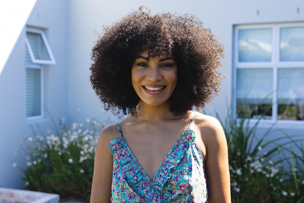 Retrato de uma linda mulher birracial sorridente, de meia idade, com cabelo afro, encostada em uma casa no quintal