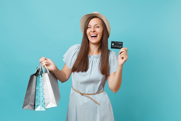 Retrato de uma linda mulher caucasiana com vestido de verão, chapéu de palha segurando sacolas de pacotes com compras após as compras, cartão de crédito do banco isolado em fundo azul pastel. Copie o espaço para anúncio.