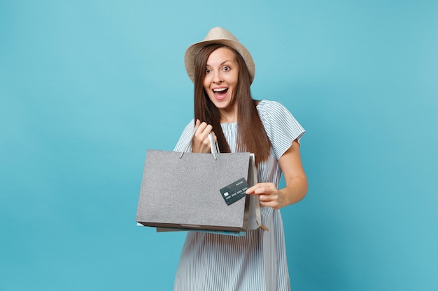 Retrato de uma linda mulher caucasiana com vestido de verão, chapéu de palha segurando sacolas de pacotes com compras após as compras, cartão de crédito do banco isolado em fundo azul pastel. copie o espaço para anúncio.