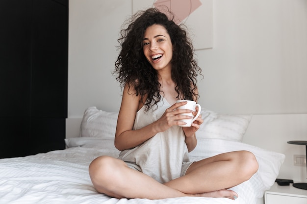Retrato de uma linda mulher feliz com cabelo longo cacheado, sentado na cama com roupa branca limpa em casa pela manhã, e segurando uma xícara de chá