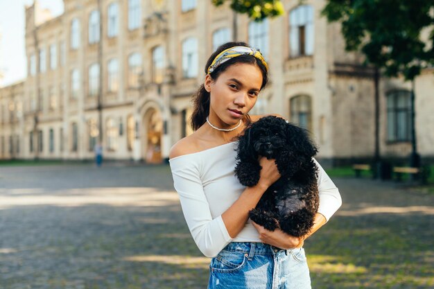 Retrato de uma linda mulher hispânica com um cachorro preto fofo nos braços em pé na rua