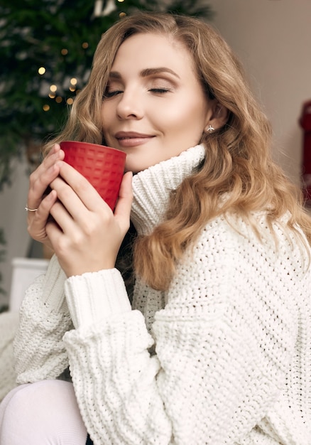 Retrato de uma linda mulher loira com um suéter de lã branco tomando café quente em um ambiente aconchegante e leve