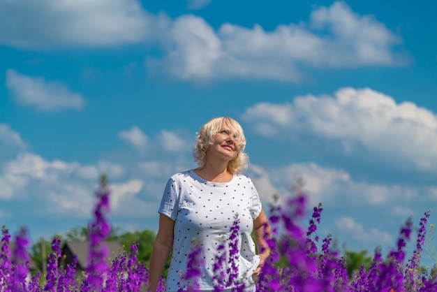 Retrato de uma linda mulher loira de meia-idade feliz em um campo com flores, fundo de céu azul