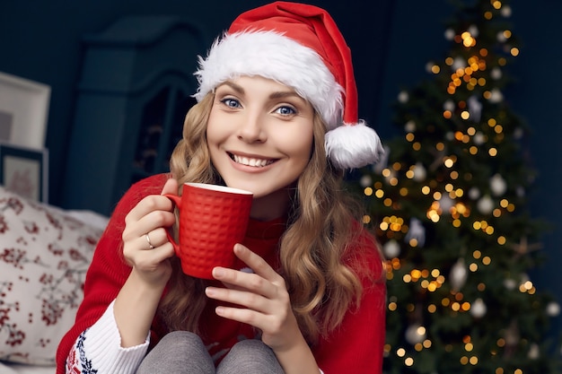 Retrato de uma linda mulher loira de suéter vermelho e chapéu de papai noel com xícara de café, posando no interior decorado de natal.