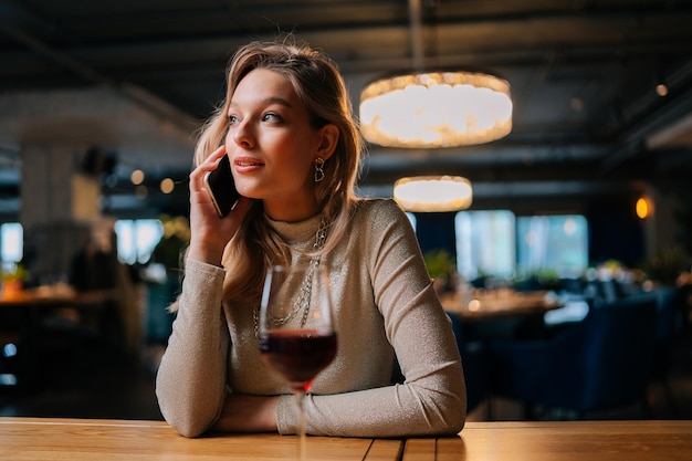 Retrato de uma linda mulher loira em roupas da moda falando no smartphone com o namorado sentado na mesa do restaurante com um copo de vinho tinto