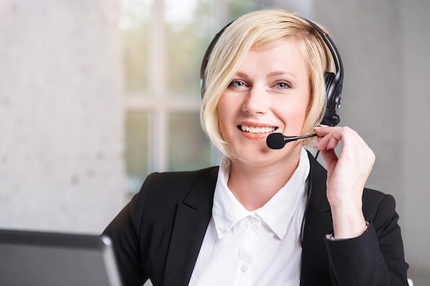Retrato de uma linda mulher loira sorridente, operadora de call center vestida de terno preto elegante com fones de ouvido trabalhando no escritório branco
