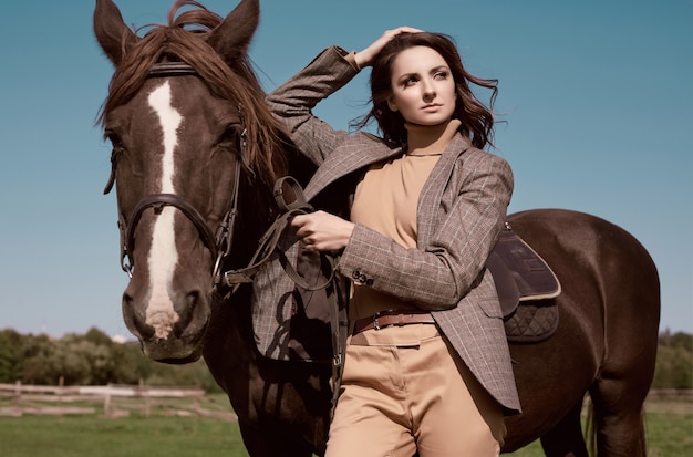 Foto retrato de uma linda mulher morena com uma elegante jaqueta xadrez marrom posando com um cavalo na paisagem do campo