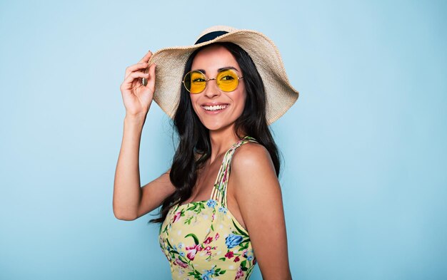 Retrato de uma linda mulher morena de verão em óculos escuros de chapéu e uma garota estilosa de vestido colorido se divertindo e posando em fundo azul