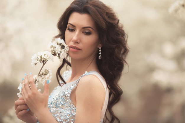 Retrato de uma linda mulher morena encaracolada em vestido de noite no jardim de árvores de flores